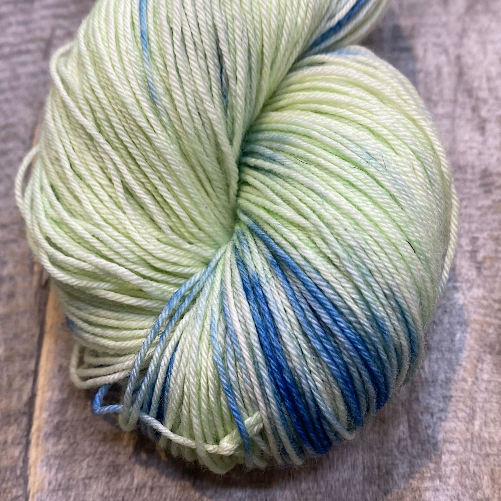 A close-up photo of the end of a skein of hand dyed wool in colour, 'East Beach'. A pale green/yellow background with blue dye painted through.