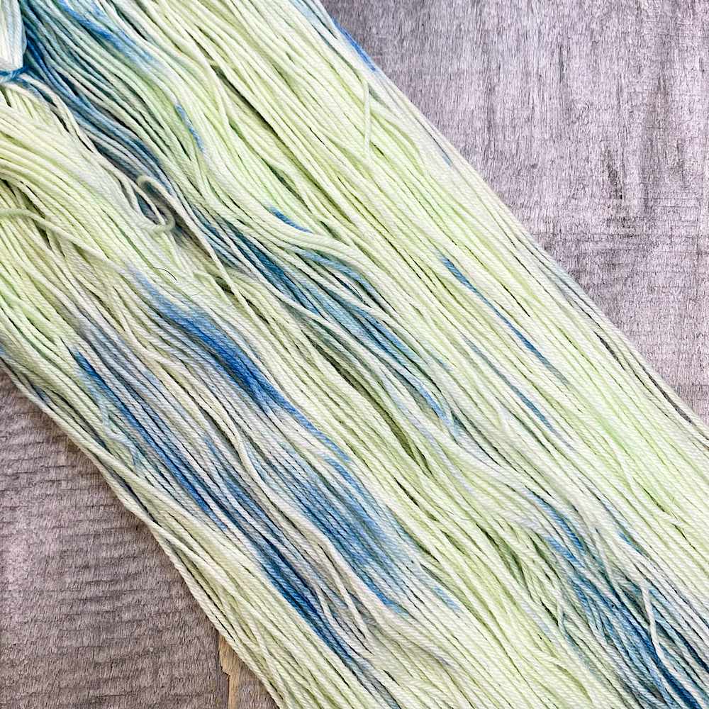 A skein of hand dyed wool in colour, 'East Beach'.. It is laid flat to show off the colours clearly - a pale green/yellow background with blue dye painted through.
