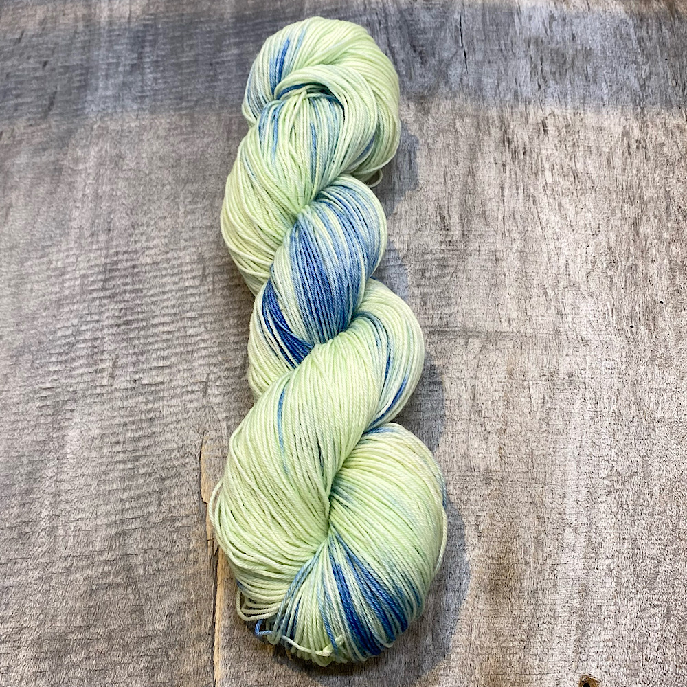 A skein of hand dyed wool in colour, 'East Beach'. A pale green/yellow background with blue dye painted through.